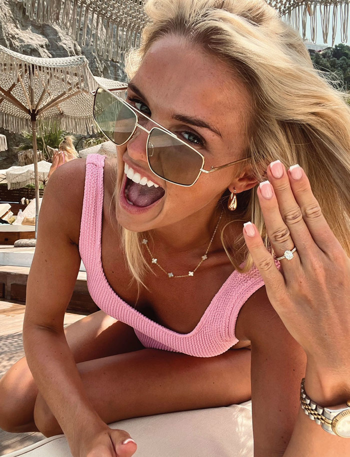 an elated woman wearing a pink swimsuit and sunglasses lifting her hand to show her diamond engagement ring