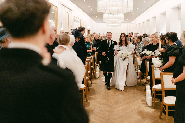 brunette bride wears high leg slit wedding dress with puff sleeves and walks down the aisle with a man dressed in a full kilt outfit