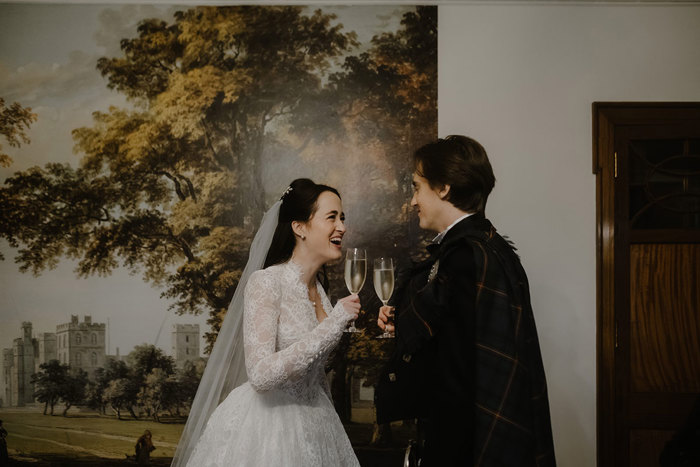 a bride and groom toasting with champagne glasses. They are standing in front of a painted mural with trees and a castle.