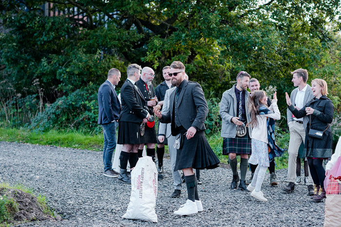 A Man Wearing A Black Kilt Stands On A Stone Path Behind A Bag Of Potatos While Holding A Potato And Other People Stand In Background