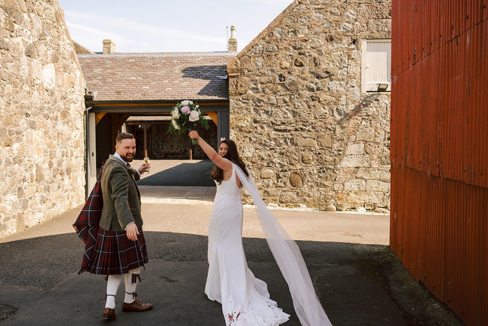 A bride and groom walking away from the camera outside, they turn back to look at the camera with a hand up in celebration