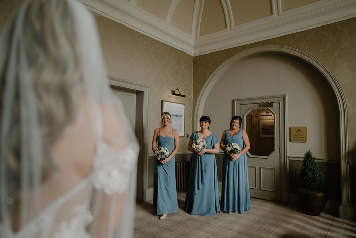 three bridesmaids wearing floor-length blue dresses smile as they look towards bride for first look at cornhill castle
