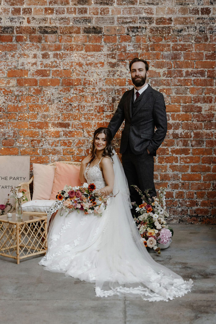 groom in grey suit stands with hand on shoulder of seated bride holding wildflower bouquet and wearing long flowy veil