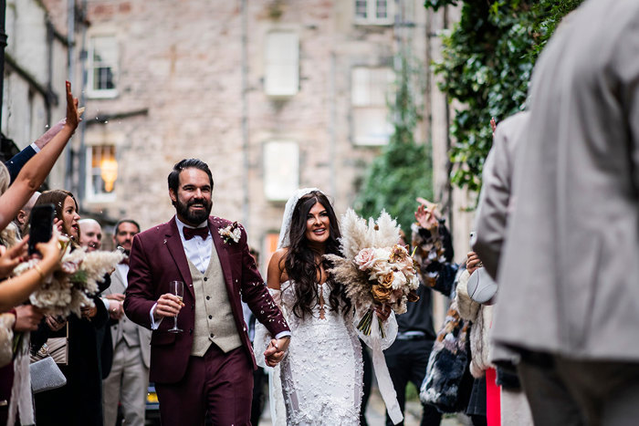 bride and groom, covered in remnants of confetti, hold hands as they walk outdoors amongst friends and family