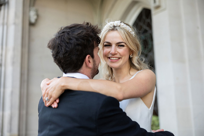 Bride smiles to the camera as she embraces groom