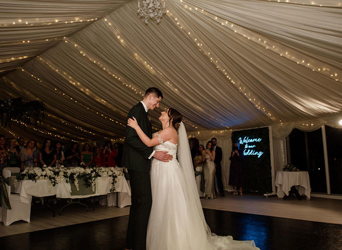 A bride and groom dance together on a dancefloor underneath fairylights in a marquee