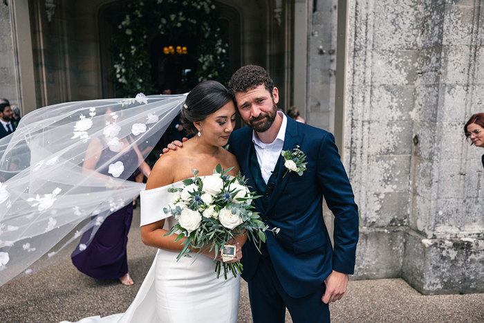 an emotional groom wraps an arm around a bride whose floral veil catches in the breeze while standing outside Blairquhan Castle