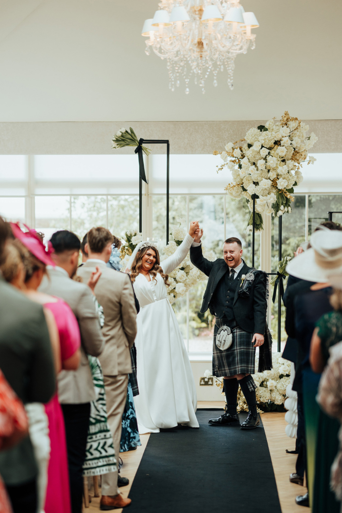 A bride and groom hold their hands up in celebration in front of their guests during their ceremony