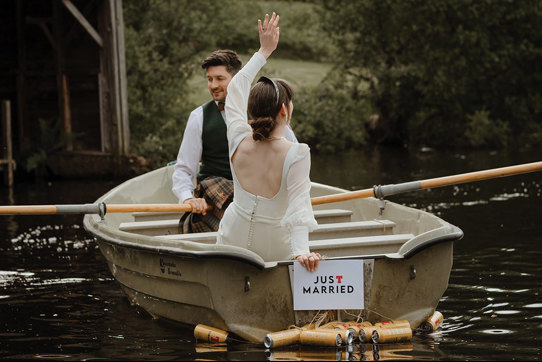 bridal couple sat in a row boat with a just married sign and beer cans hanging off the back of the boat