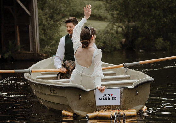 bridal couple sat in a row boat with a just married sign and beer cans hanging off the back of the boat