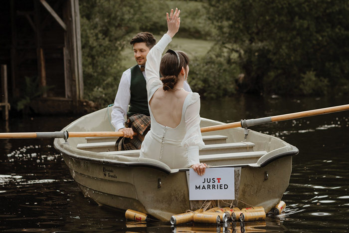 bridal couple sat in a row boat with a just married sign and beer cans hanging off the back of the boat