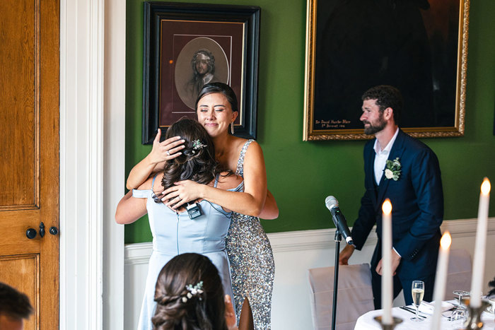 a bride wearing a silver glittery dress hugs a bridesmaid wearing a pale blue silk dress. The groom stands up from table in background