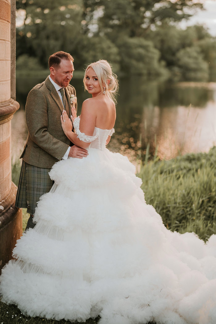 A bride wearing a wedding dress with a large skirt standing next to a groom wearing a green kilt looking over her shoulder at the camera