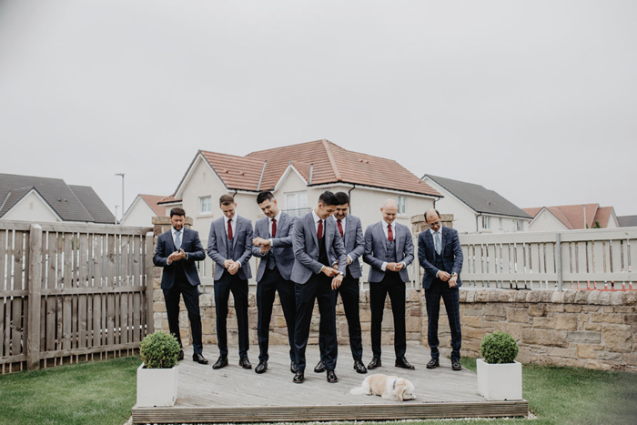 A group of men in grey and black suits with red or blue ties adjust their sleeves