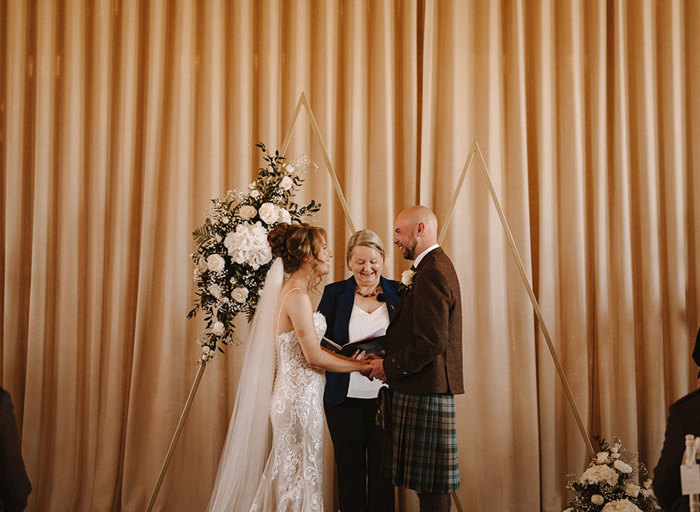 a bride and groom stand in front of a decorative triangular wooden ceremony arch as a wedding celebrant reads notes from a book.