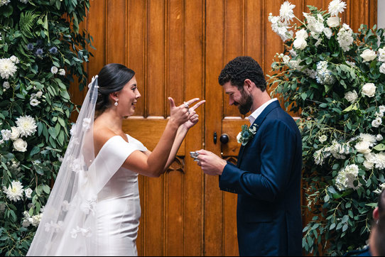 A bride makes a pointed finger gesture while a groom wearing a dark blue suit looks down at a silver quaich vessel in his hands, while standing against a large wooden door at Blairquhan Castle