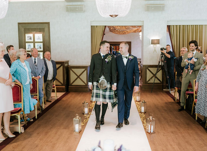 grooms holding hands walking up a white carpet aisle as guests either side clap