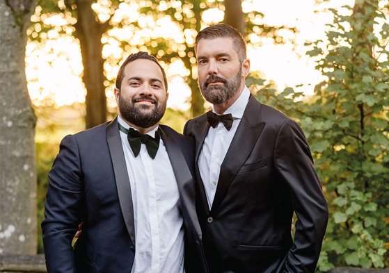 two grooms wearing suits and bowties stand with their arms around each other