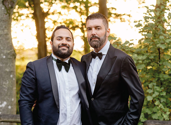 two grooms wearing suits and bowties stand with their arms around each other