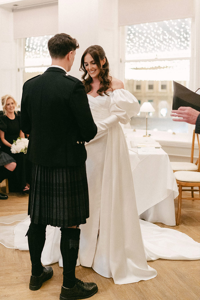 view from the back of groom in full dark tartan kilt outfit as he holds hands with smiling brunette bride in long veil and wedding dress with puff sleeves as others look on