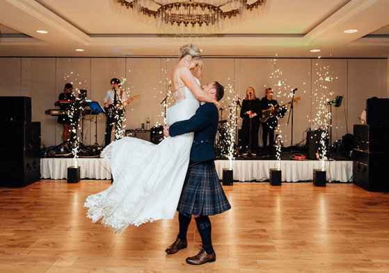 a groom lifting a bride up on a wooden floor. There is a band on a small stage behind them with pyrotechnics.