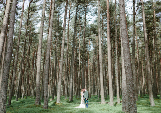 Couple in wedding outfits standing amongst tall trees in forest setting