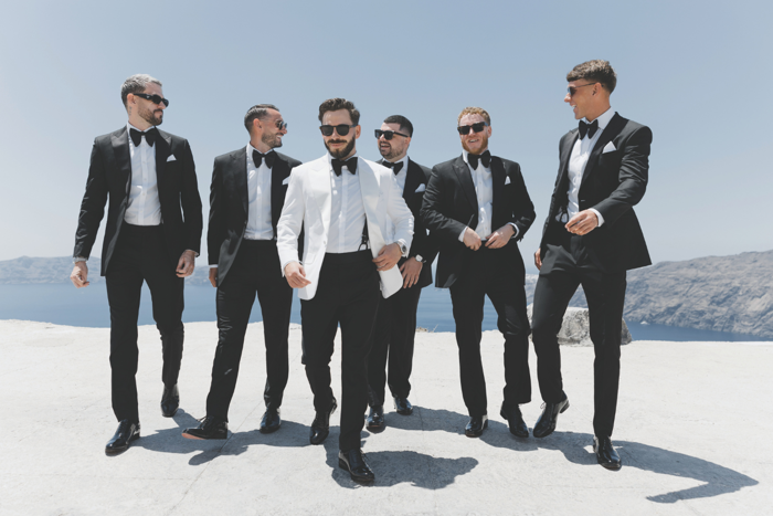 six men walking on top of cliff, five wearing black tuxedos and one wearing white tuxedo and black trousers