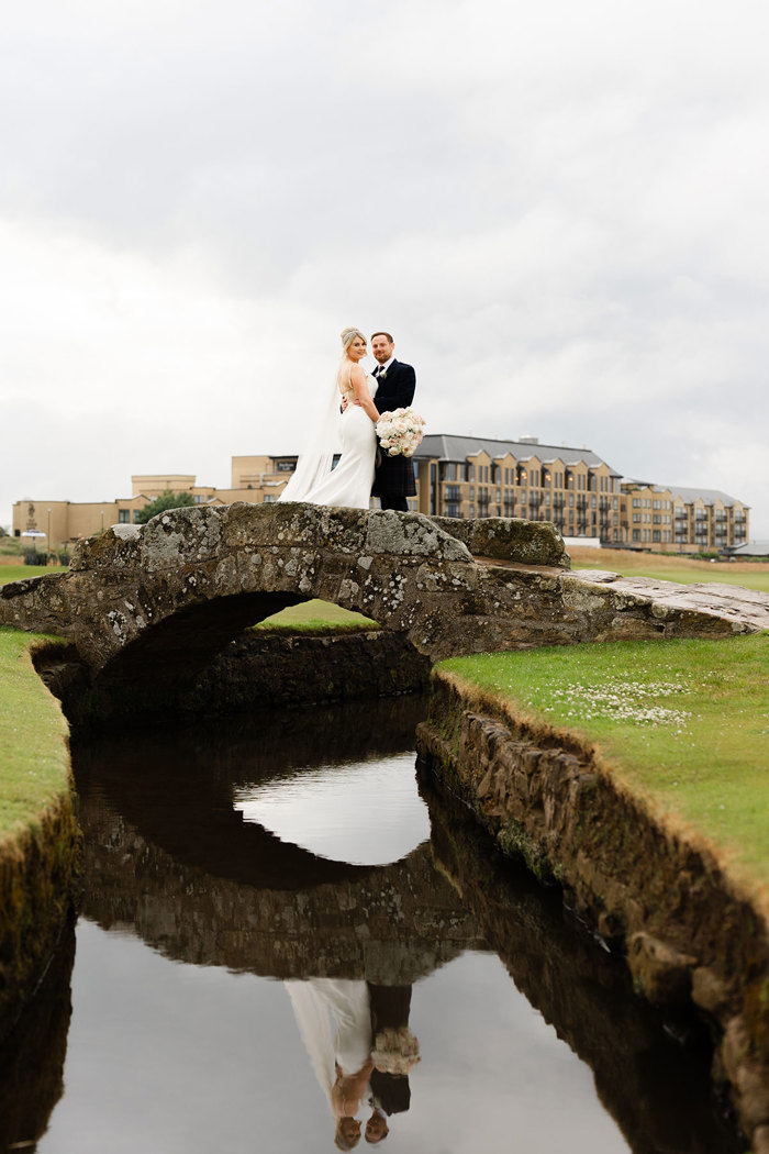 a bride and groom pose on Swilcan Bridge with Old Course Hotel in background