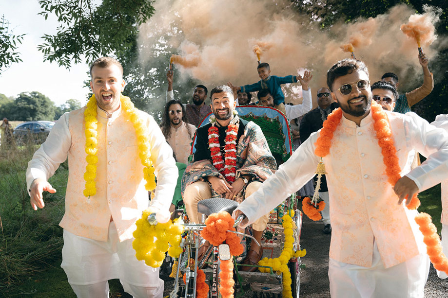 A group of men in white outfits with colourful garlands around their necks and a person riding a cart with smoke bombs.