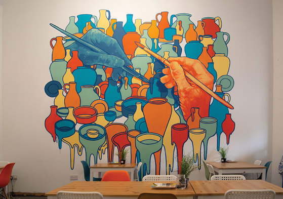 a bold coloured wall mural depicts two large hands each holding a paint brush and lots of ceramic pots and vases around them with the bottom row looking to be dripping paint