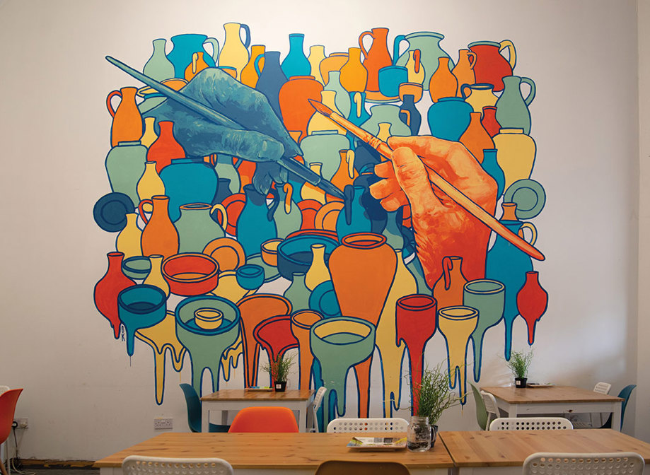 a bold coloured wall mural depicts two large hands each holding a paint brush and lots of ceramic pots and vases around them with the bottom row looking to be dripping paint