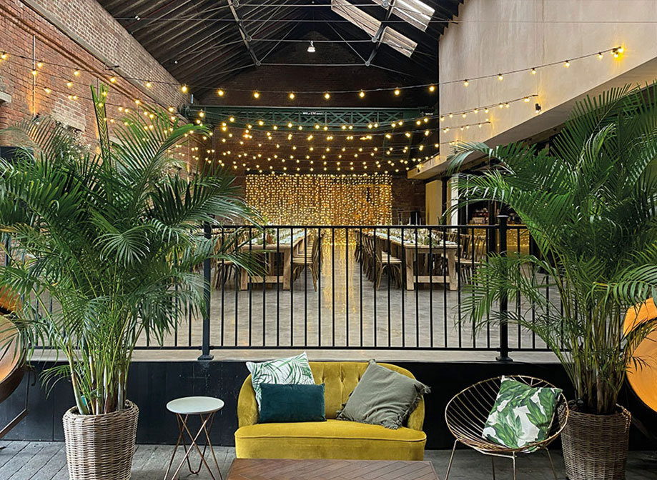 An industrial room with long wooden tables and fairylights draping the walls in the background and a yellow couch with plants on either side in the foreground