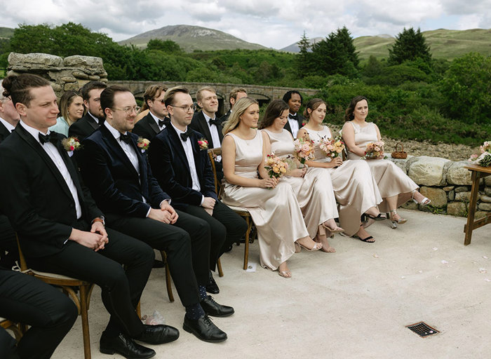 a row of wedding guests wearing smart attire watching a wedding ceremony outside