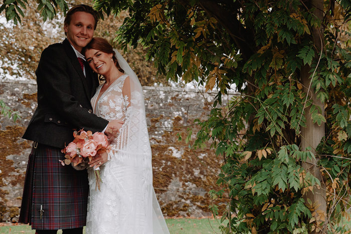 bride and groom both smile as they stand outside surrounded by autumnal foliage and lean into each other, the bride resting her head on the groom's lower shoulder