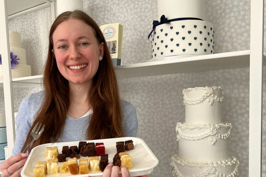a brown haired woman stands in front of shelves with wedding cakes holding a tray of tiny different flavoured cake slices