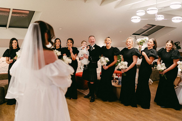 brunette bride stands at forefront as nine bridesmaids in black dresses and a man holding a little flowergirl all smile and look excited