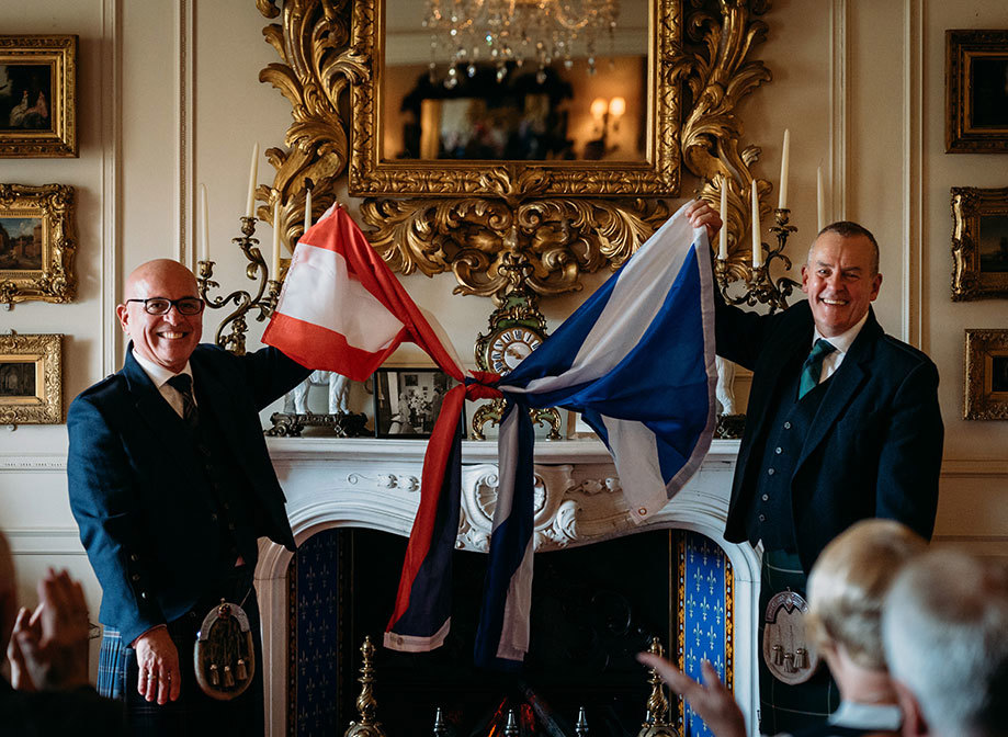 two grooms standing in front of a fireplace and below a gilded mirror hold up Scottish and Puerto Rican flag knotted together