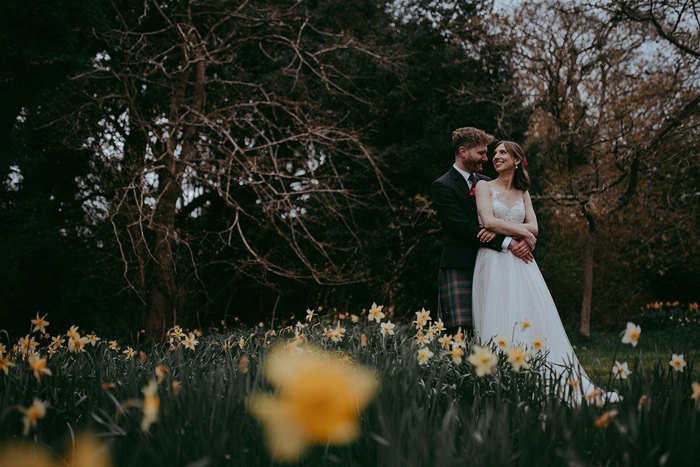 a smiling bride and groom standing in a field of yellow daffodils with trees behind them