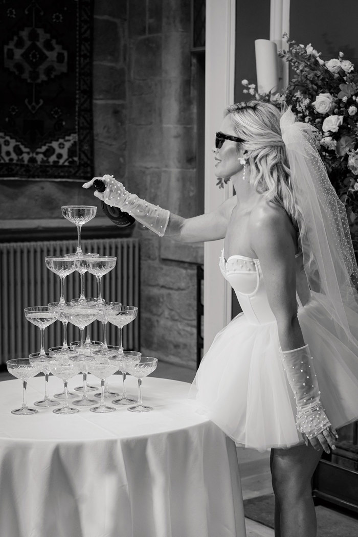 A bride wearing a short dress with tulle sleeves and a tulle bow and sunglasses pours prosecco into a tower of coupe glasses