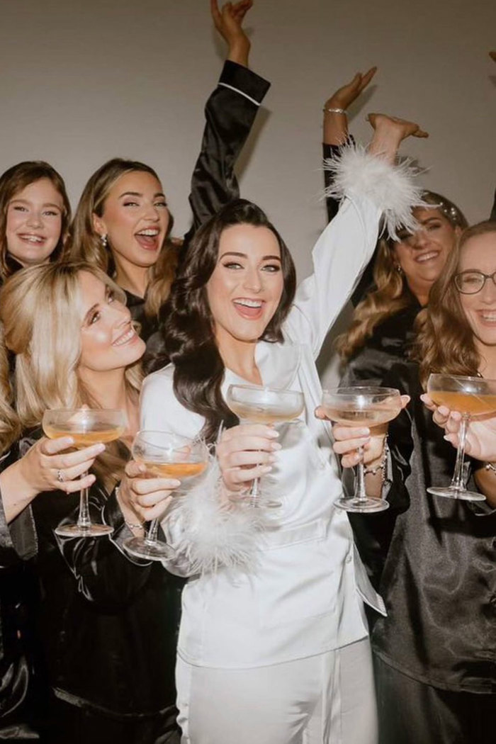 bride in white fluffy pajamas raising cocktail glasses in the air alongside other women with cocktails wearing black fluffy pajamas