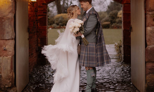blonde bride in feathery cape and white wedding dress holds bouquet of white roses and stands smiling and facing groom in green jacket and red, blue, green kilt with matching fly plaid, both stood under outdoor stone archway