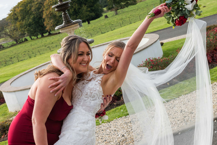 A Bride Cheering And Waving Her Bouquet In The Air Being Held By A Person Wearing A Red Dress