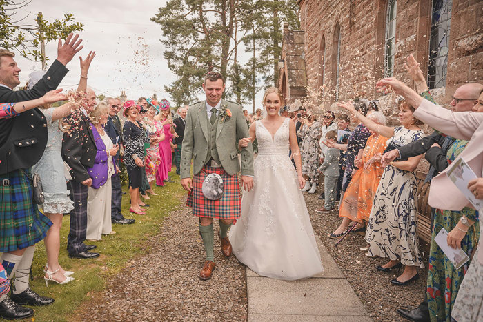 A bride and groom walk hand in hand along a path as their guests on either side throw confetti