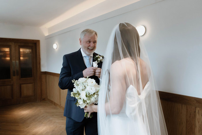 A bride wearing a veil and a large bow on the back of her dress faces away from the camera as she does a first look with her father who is smiling