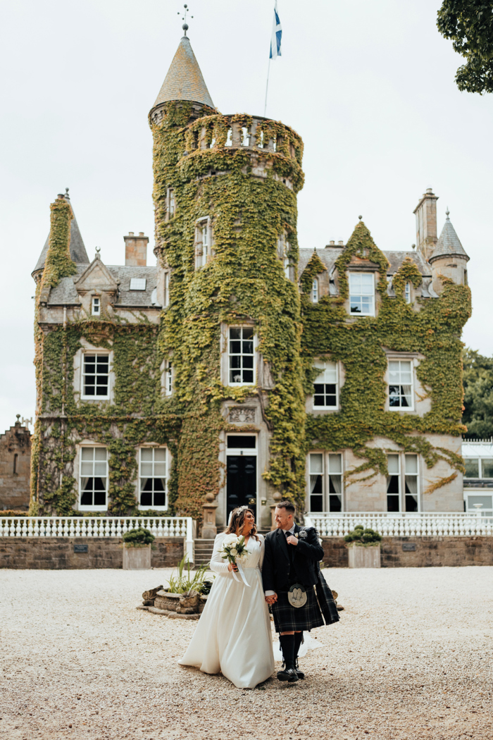 A man in a dark kilt and a bride in a long sleeved wedding dress walk towards the camera away from a castle covered in greenery