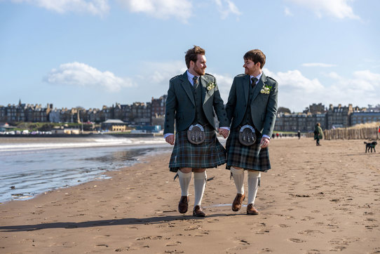 two grooms walking hand in hand along a sandy beach with St Andrews skyline in the background