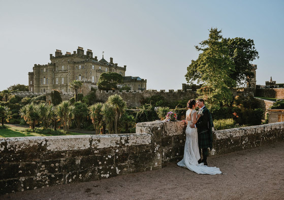 A bride and groom kissing standing by a wall overlooking Culzean Castle and its gardens