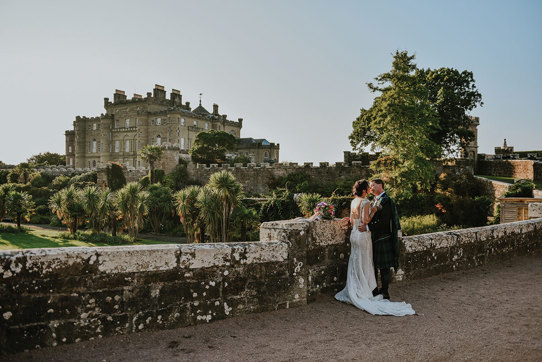 A bride and groom kissing standing by a wall overlooking Culzean Castle and its gardens