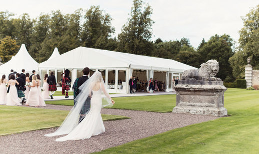 Bride, groom, wedding party and bag pipers walk across pebble path to get to white 