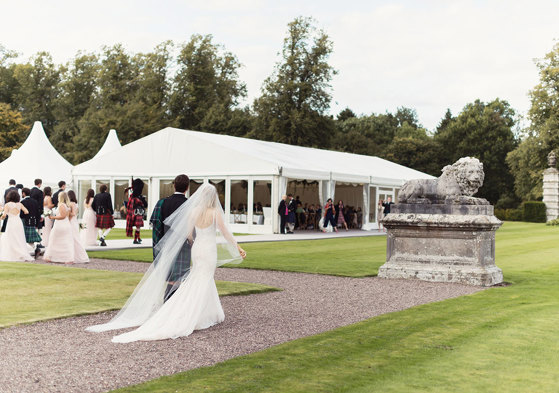 Bride, groom, wedding party and bag pipers walk across pebble path to get to white 